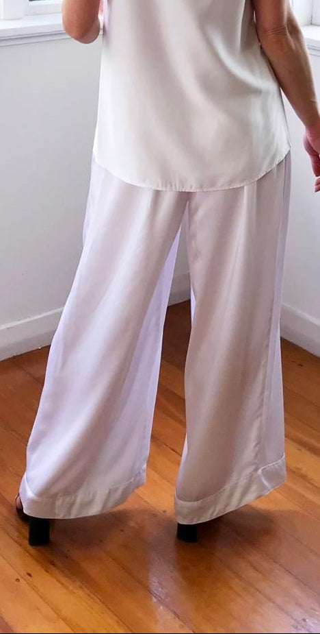 Woman wearing a white sleeveless top and light gray pants standing in a room with large windows.
