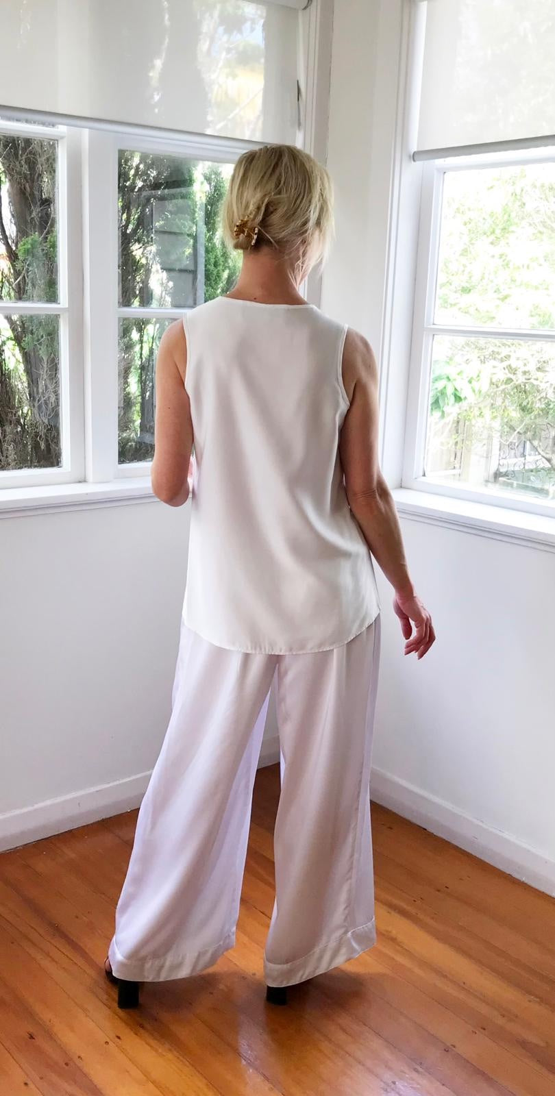 Woman wearing a white sleeveless top and light-colored pants standing in a room with large windows.