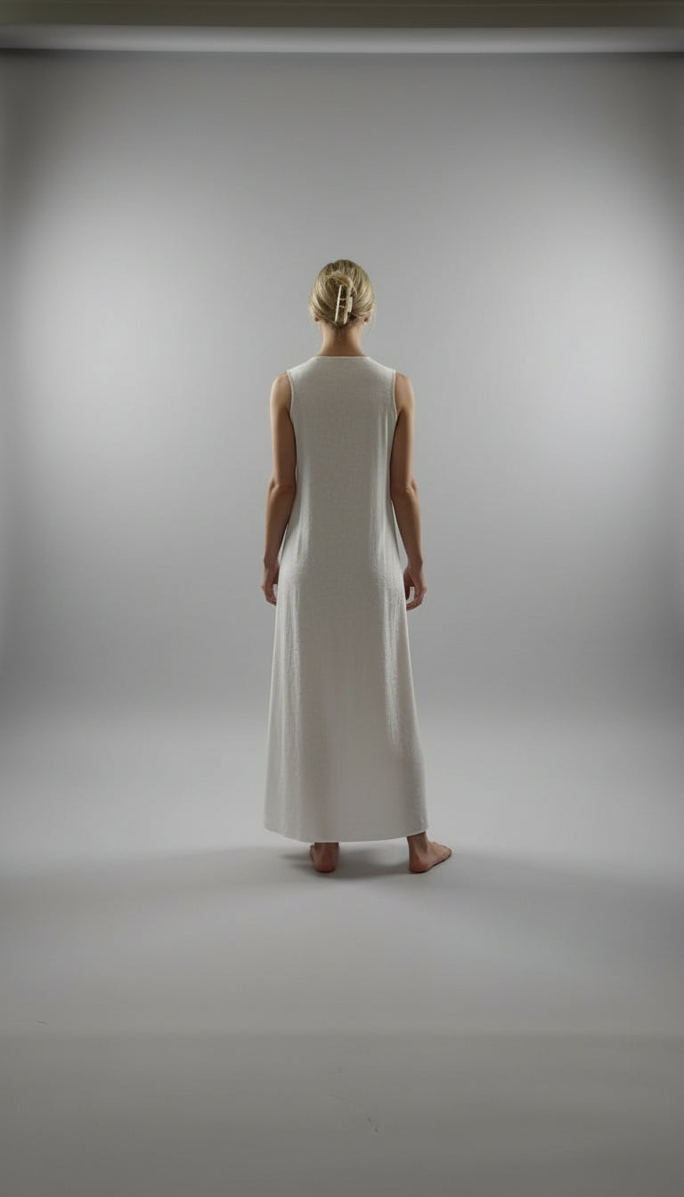 Woman wearing a white dress standing in a room with wooden floor and large windows.