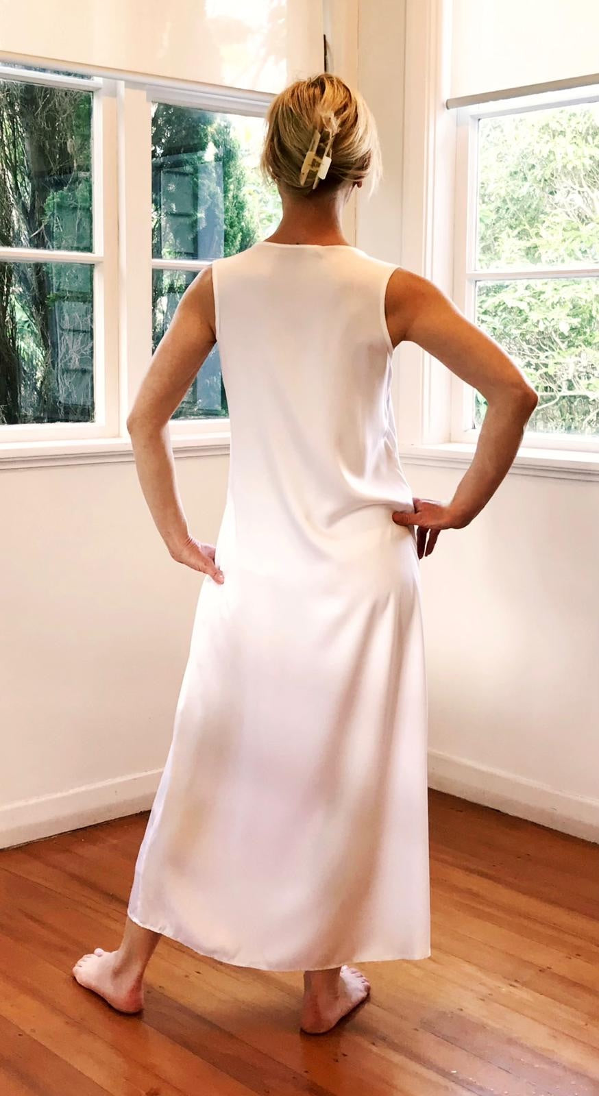 Person wearing a white sleeveless dress standing in a room with large windows.