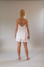 Person wearing a white slip dress standing in a room with wooden flooring and large windows.