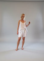 Woman wearing a white slip pyjamas standing in a room with wooden flooring and large windows.