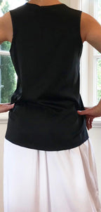 Person wearing a black sleeveless top and white pants standing indoors with large windows in the background.