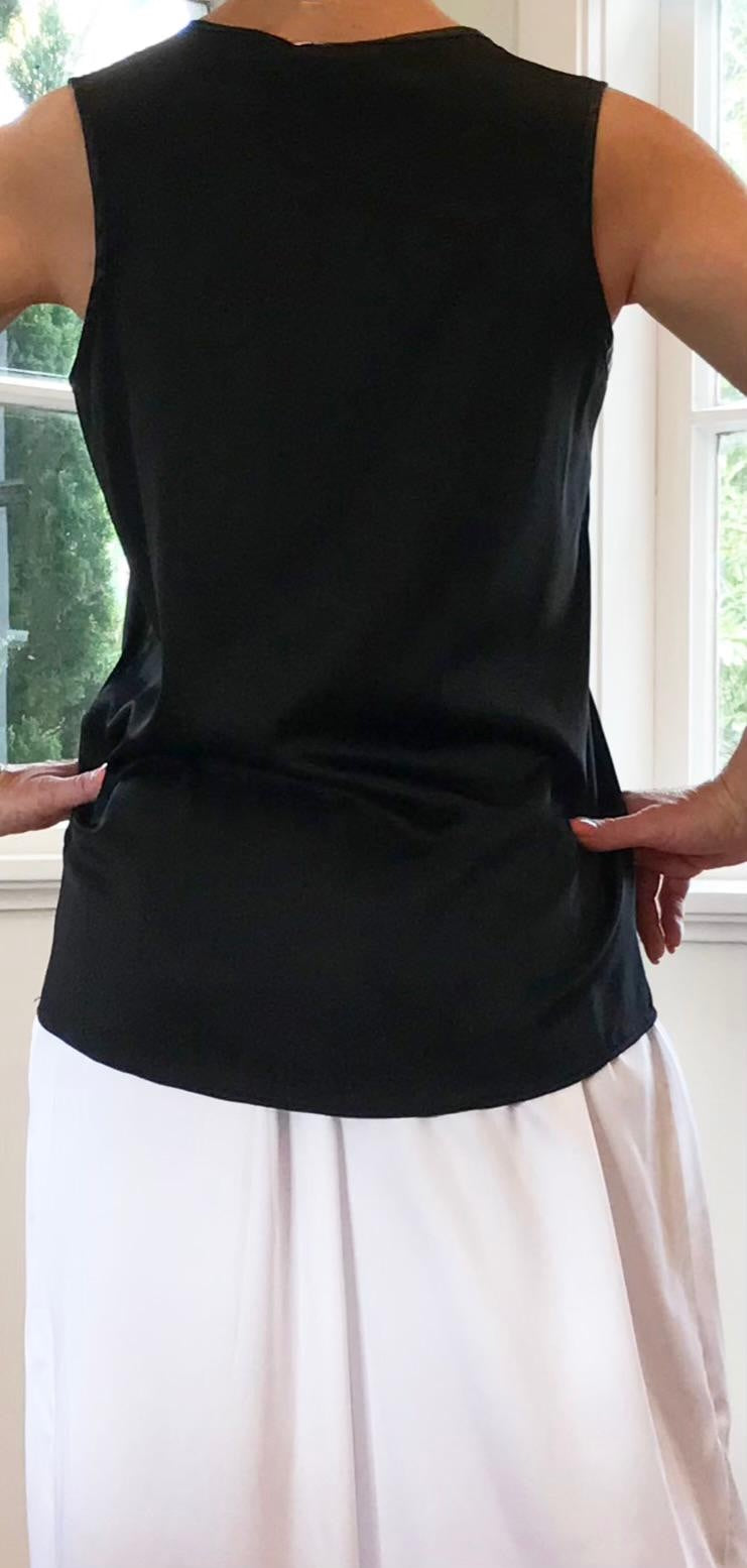 Person wearing a black sleeveless top and white pants standing indoors with large windows in the background.